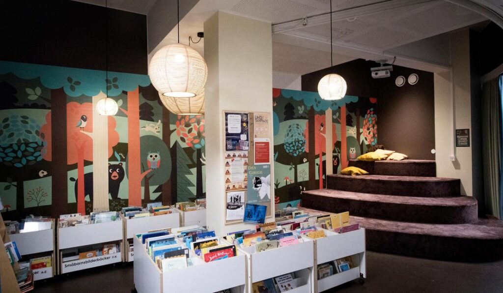 Barnavdelningen i Hammarkullens bibliotek med bokbackar, en målad skogsvägg och en trappläktare med kuddar.